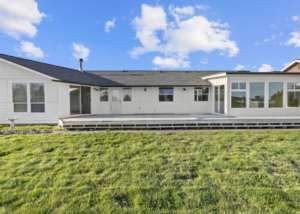 White ranch-style house with a long wooden deck and large windows, under a bright blue sky and green lawn.