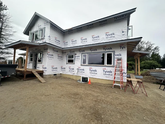 home-service-custom-homes | Laika Construction | Bellingham Contractor Two-story house under construction wrapped in Tyvek with a wooden porch and ladder on the side, on a dirt lot.